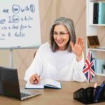 A smiling female tutor waving at a laptop during a virtual lesson with a whiteboard showing grammar rules and a UK flag on the desk, providing high-quality English tutoring 5 in Greenwich