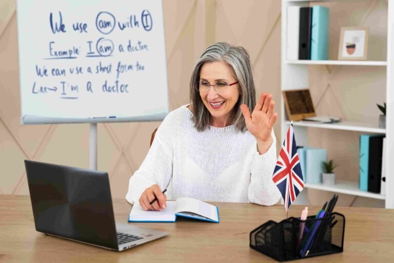 A smiling female tutor waving at a laptop during a virtual lesson with a whiteboard showing grammar rules and a UK flag on the desk, providing high-quality English tutoring 5 in Greenwich