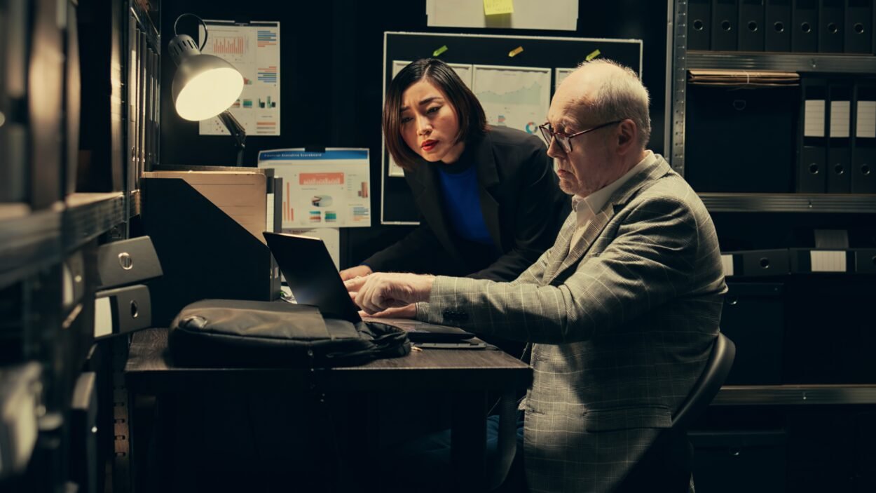 Two investigators reviewing legal records and digital files in a dimly lit office with archive shelves.