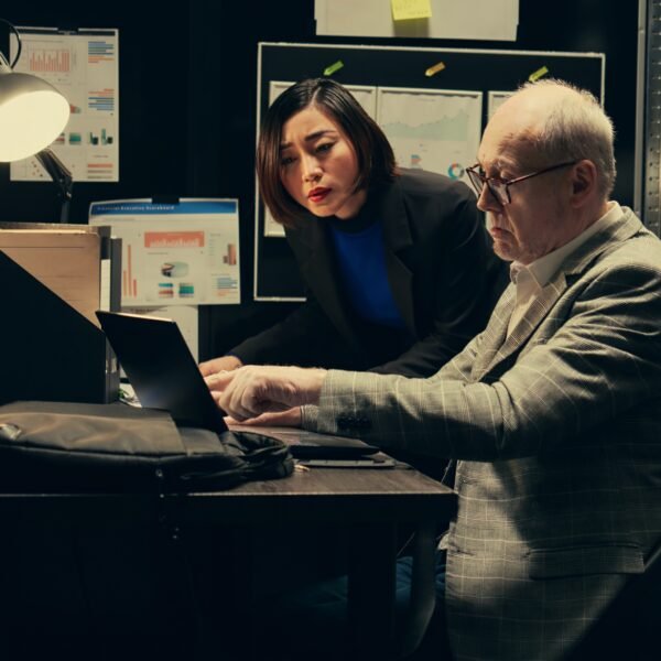 Two investigators reviewing legal records and digital files in a dimly lit office with archive shelves.